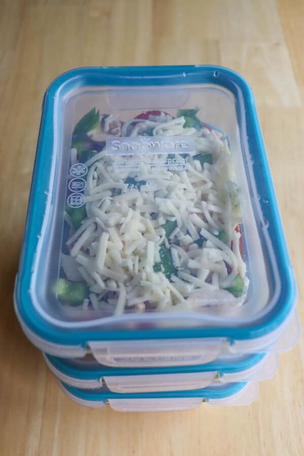 A stack of three rectangular, clear plastic containers with blue lids rests on a wooden surface. The top container resembles a cottage cheese pizza bowl, bursting with shredded cheese atop a colorful bed of mixed vegetables.