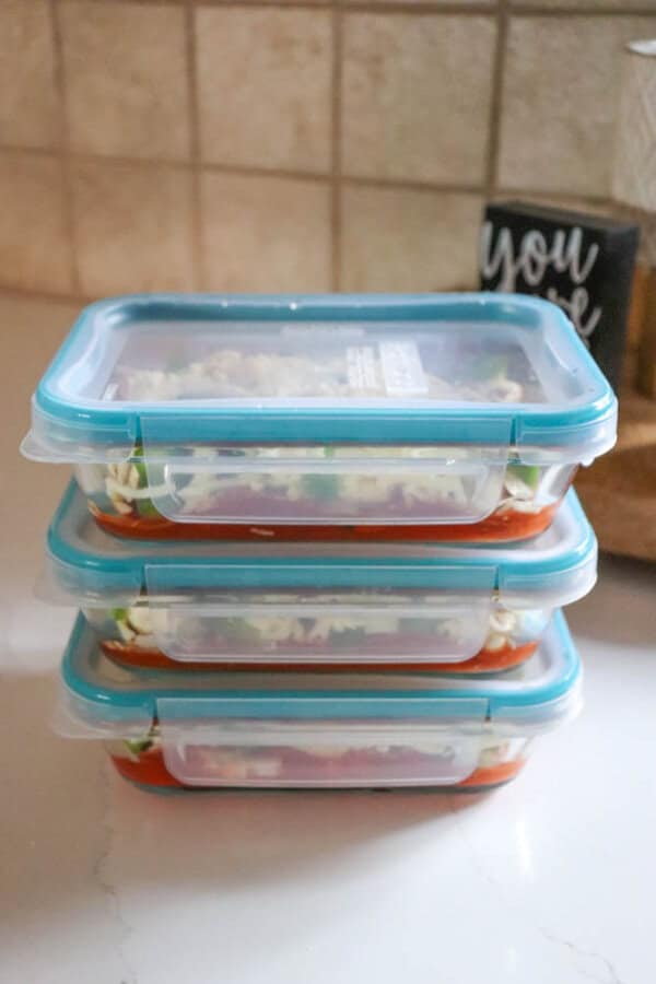 Three stacked, clear food containers with blue lids sit on a countertop. The containers appear to hold layered meals, possibly cottage cheese pasta dishes with visible cheese and sauce. A small decorative sign is partially visible in the background.