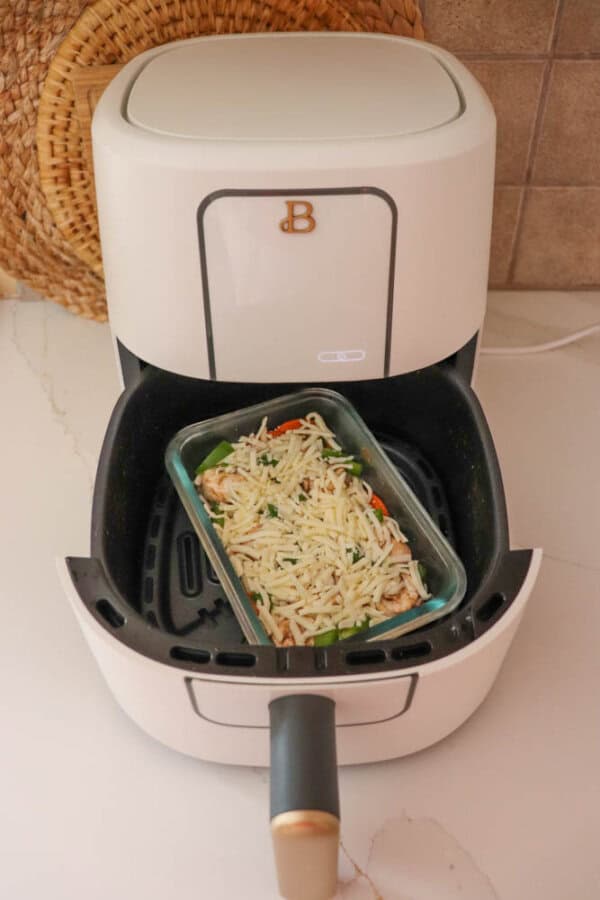 An air fryer with a glass dish inside, featuring a blend of chopped vegetables and grated cheese reminiscent of cottage cheese pizza bowls. The fryer, white with a sleek black interior, rests on a light-colored countertop beside a charming woven basket.