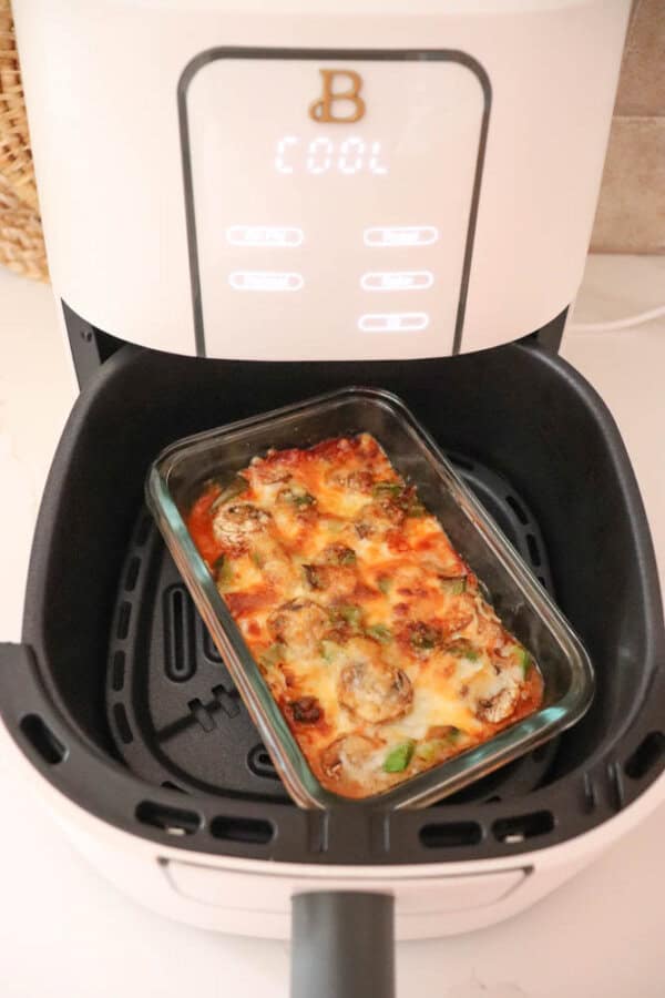 A glass dish with baked vegetables and melted cheese, reminiscent of a delightful cottage cheese pizza bowl, rests inside an air fryer. The air fryers display shows the word COOL, with its illuminated control buttons casting a soft glow.