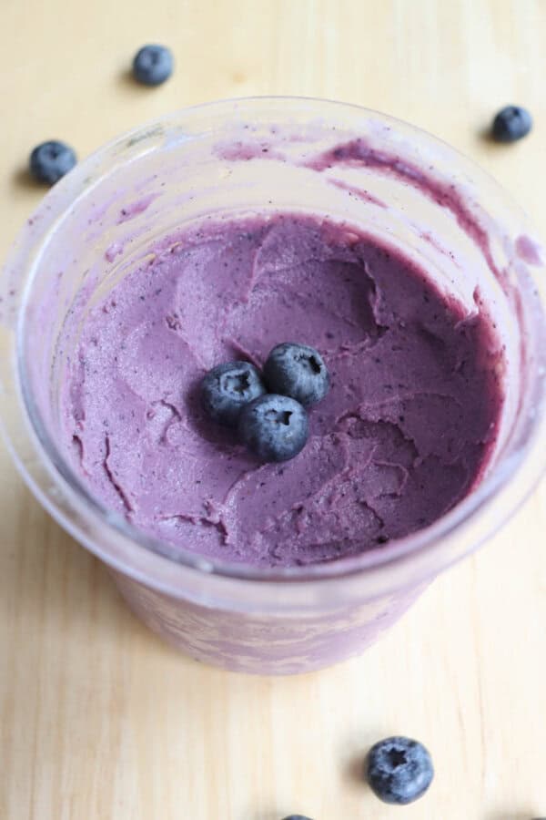 A container filled with smooth, purple blueberry frozen yogurt, topped with three fresh blueberries. A few additional blueberries are scattered on the light wooden surface around the container.
