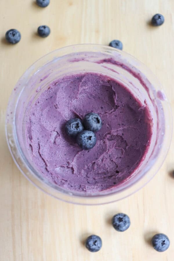 A glass bowl filled with vibrant purple blueberry mousse, topped with three fresh blueberries in the center. More blueberries are scattered on the light wood surface around the bowl.
