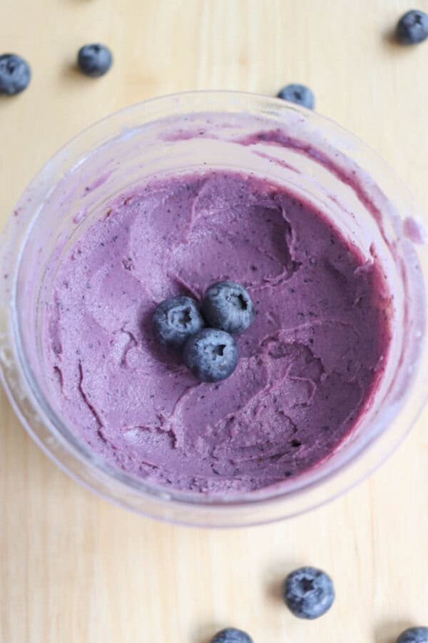 A bowl of creamy, purple blueberry frozen yogurt garnished with three fresh blueberries in the center. Several additional blueberries are scattered on the light wooden surface around the bowl.
