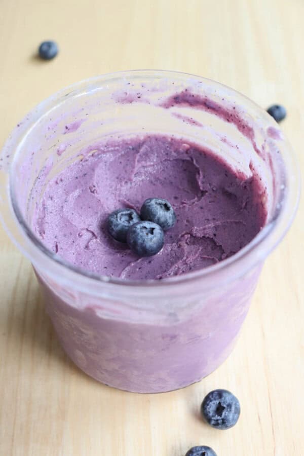 A clear container filled with purple blueberry frozen yogurt is topped with three blueberries. A few blueberries are scattered on the light wooden surface around the container.