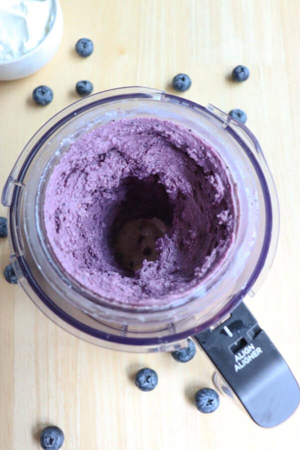 A ninja creami cup filled with purple blueberry mixture sits on a wooden surface. Blueberries are scattered around the processor, and a small bowl of yogurt is partially visible in the background.