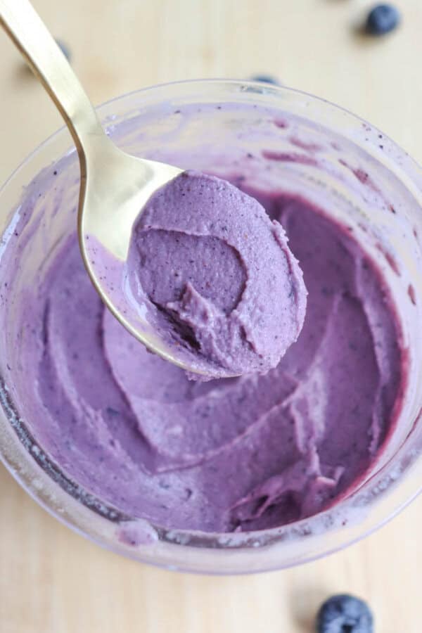 A spoonful of creamy, purple blueberry yogurt hovers above a clear bowl filled with more of the yogurt. Blueberries are scattered in the background on a light wooden surface.