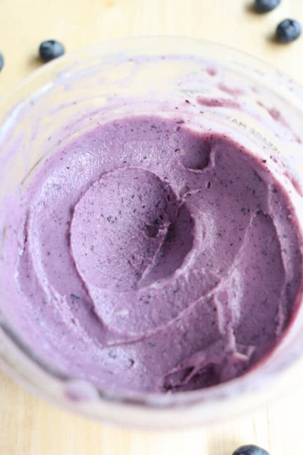A close-up of smooth, creamy purple blueberry ice cream in a bowl. The ice cream has a swirled texture, and a few blueberries are scattered on the light-colored wooden surface around the bowl.