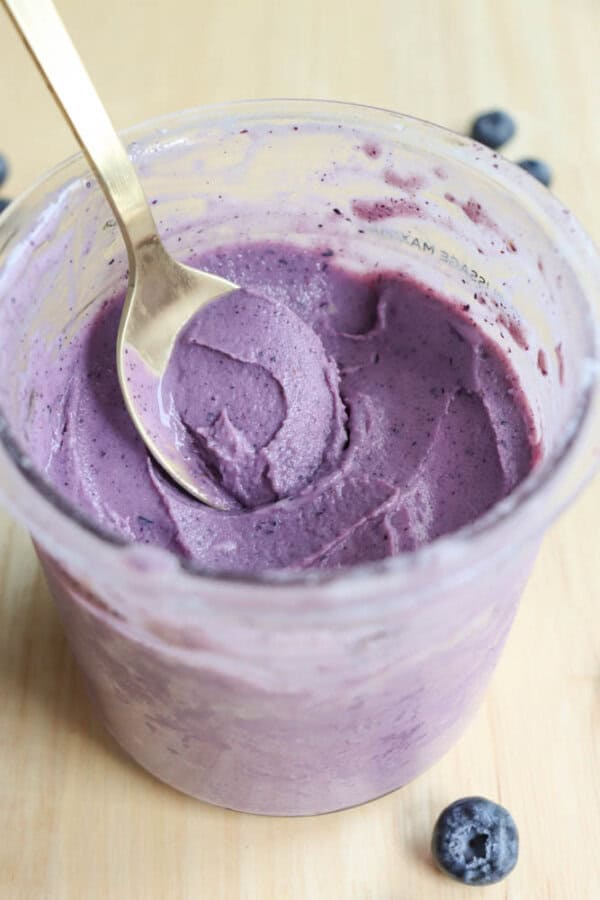 A jar of creamy purple blueberry frozen yogurt with a gold spoon inside. A few whole blueberries are scattered on the light wooden surface around the jar.