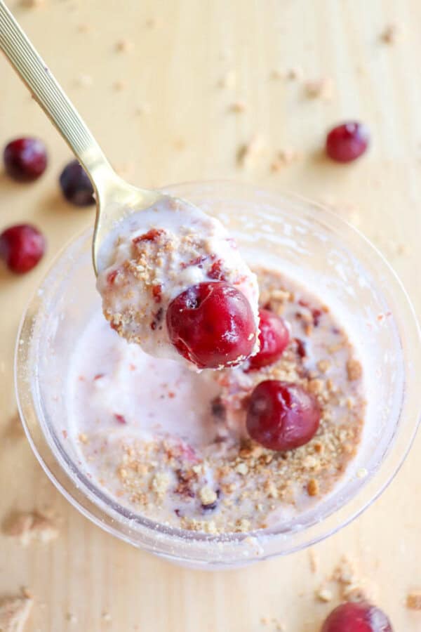 A spoonful of creamy dessert, reminiscent of a raspberry cheesecake, with cherries and crushed cookies hovers above a bowl. The bowl brims with more cherries and crumbles on a light wooden surface, surrounded by scattered cherries in the background.