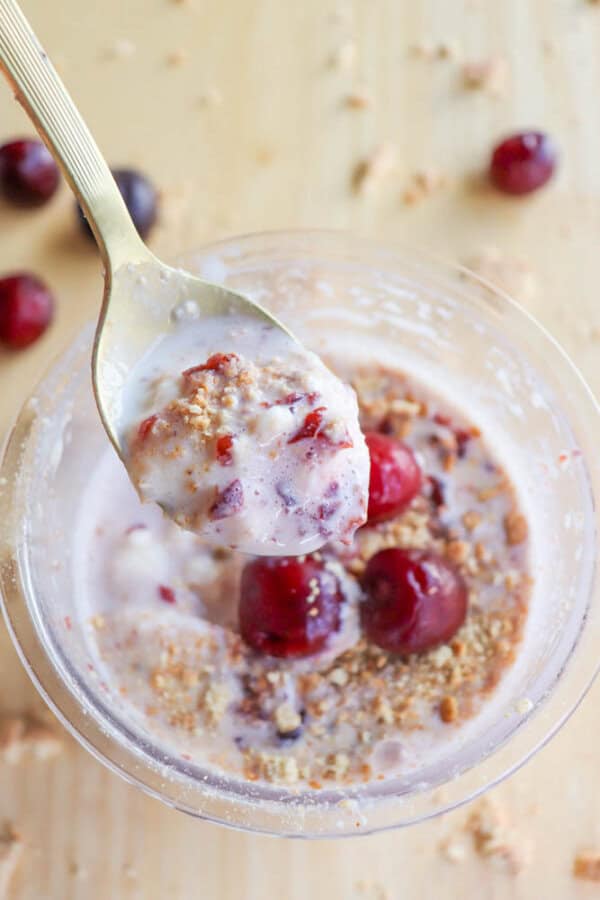 A spoonful of creamy dessert with visible cherry pieces and crushed biscuit, held above a bowl. The bowl, contains cherries, crumbly bits, and creamy mixture. A few cherries are scattered on the wooden surface.