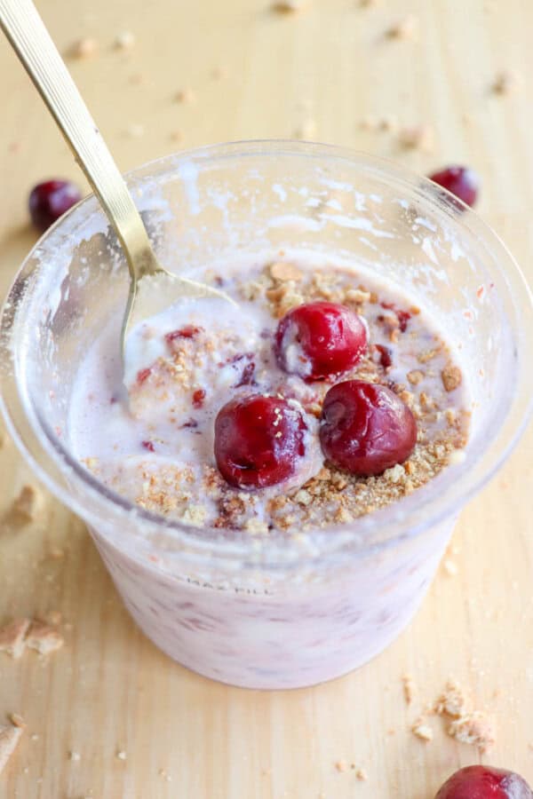 A clear pint brimming with ice cream is crowned with three cherries and crushed graham crackers, reminiscent of a cherry cheesecake delight. A spoon stands ready in the yogurt, surrounded by scattered cherries and crumbs on the wooden surface, embodying rustic charm.