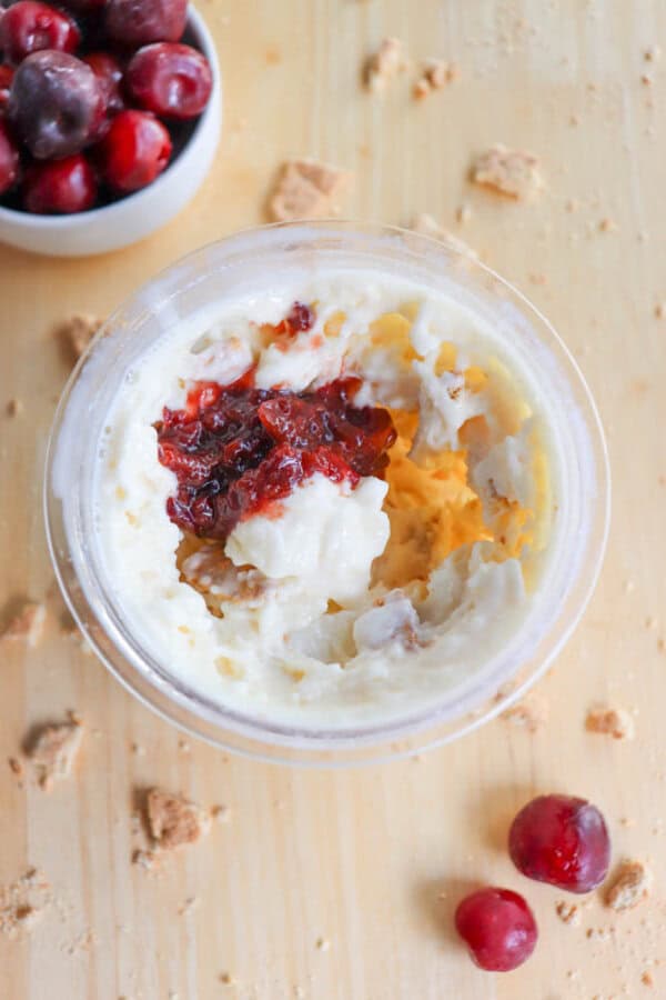 A top view of a dessert cup with creamy cheesecake filling swirled with a layer of raspberry sauce. Surrounding the cup are scattered graham cracker crumbs and a few fresh cherries. A small white bowl of cherries sits nearby, hinting at a delightful ninja creami recipe on the wooden surface.