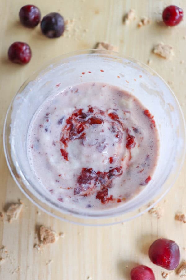 A bowl of ice cream features swirls of purple and red, hinting at berry flavors reminiscent of a raspberry cheesecake. Whole and halved cherries are scattered on the light wooden surface around the bowl, along with a few crumbs.