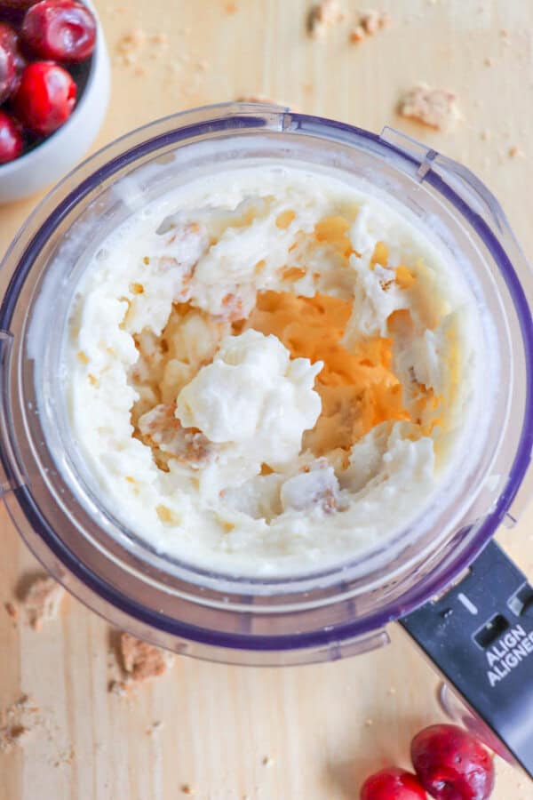 A top-down view of a food processor blending a creamy raspberry cheesecake dessert with chunks visible, perhaps ice cream or frozen yogurt. A bowl of cranberries is partially visible in the upper left corner on a wooden surface, perfect for recreating this Ninja Creami recipe.
