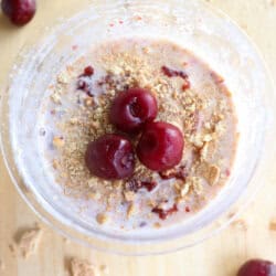 A plastic pint of cherry cheesecake ice cream topped with three cherries and crushed graham crackers. Additional cherries are scattered on the light wooden surface around the bowl, creating a ninja creami recipe masterpiece.