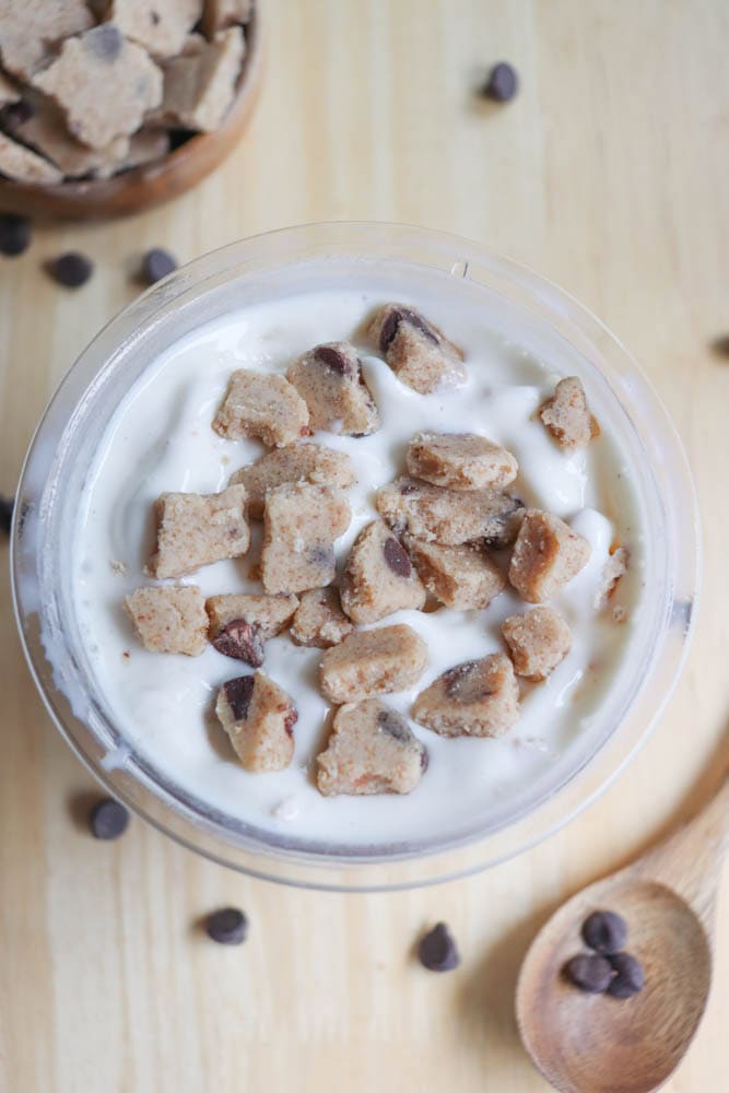 A bowl of creamy yogurt is topped with chunks of chocolate chip cookie dough. Nearby, a wooden spoon and scattered chocolate chips add to the inviting presentation. A small bowl of additional cookie dough pieces is also visible in the background.