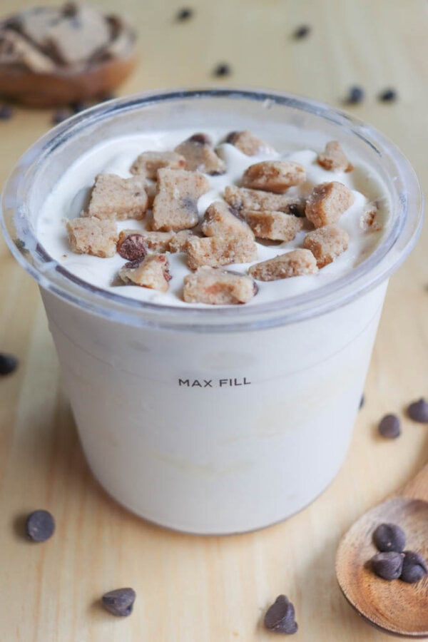 A cup of vanilla ice cream filled to the max fill line is topped with cookie dough pieces and chocolate chips. A few chocolate chips are scattered on the wooden surface, with a wooden spoon lying nearby.