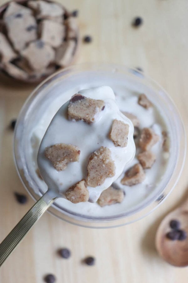 A spoonful of creamy vanilla ice cream mixed with chunks of cookie dough is being held above a bowl. In the background, a wooden bowl and a spoon are filled with additional cookie dough pieces, and a few chocolate chips are scattered on the surface.