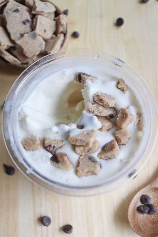 A bowl of vanilla ice cream with chunks of chocolate chip cookie dough on top. Some cookie dough pieces are scattered around the bowl on a wooden surface. A wooden spoon and additional chocolate chips are nearby.