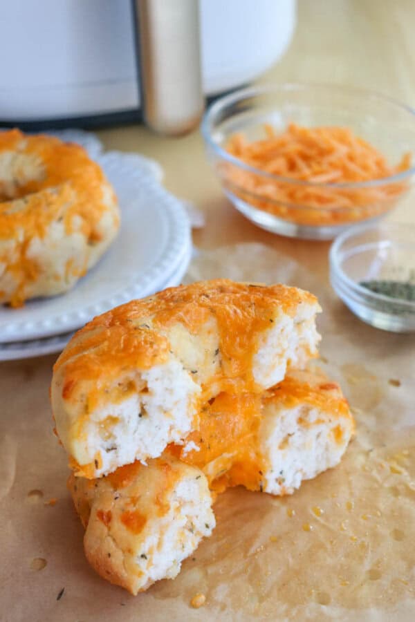Cheese bagels are stacked on parchment paper, one with a bite taken out. Behind them, theres a small bowl of shredded cheese and another with herbs. A plate holds more biscuits. The background features a kitchen appliance.