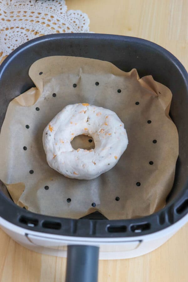 An uncooked bagel with visible orange flecks rests on parchment paper inside an air fryer basket. The parchment is perforated, and a lace doily is partially visible on the wooden table surface.