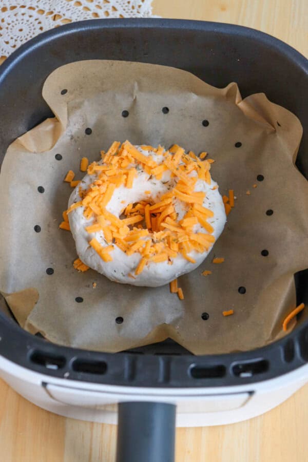 A bagel topped with shredded cheddar cheese is placed on a piece of parchment paper inside an air fryer basket. The basket sits on a wooden surface, and a corner of lace fabric is seen in the background.