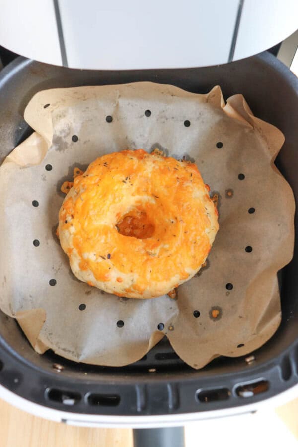 A cheese bagel sits on parchment paper inside an air fryer. The melted cheese topping has browned slightly, suggesting it has been freshly baked.