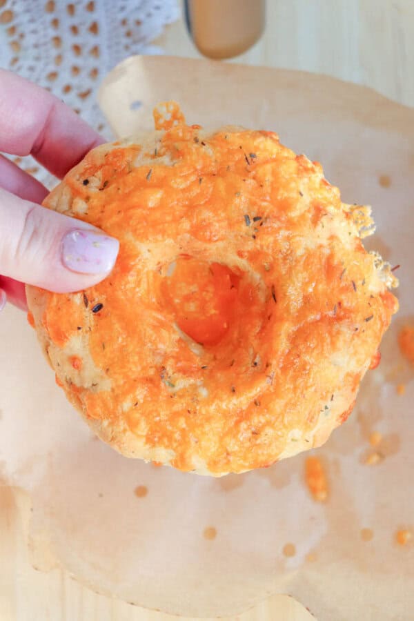 A hand holding a cheese bagel with melted cheese and herbs on top, above a piece of parchment paper. Theres a textured, golden-brown crust visible in the sunlight.