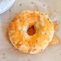 A freshly baked cheddar cheese bagel sits on parchment paper, showcasing a golden-brown crust with melted cheese on top. Theres a glimpse of another bagel on a white plate in the corner.