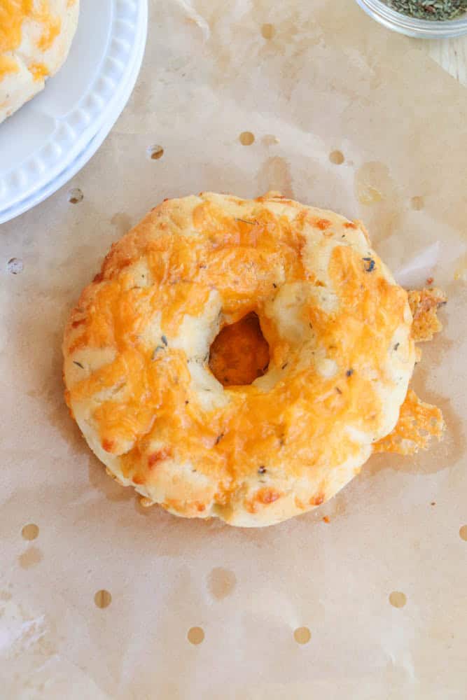 A freshly baked cheddar cheese bagel sits on parchment paper, showcasing a golden-brown crust with melted cheese on top. Theres a glimpse of another bagel on a white plate in the corner.