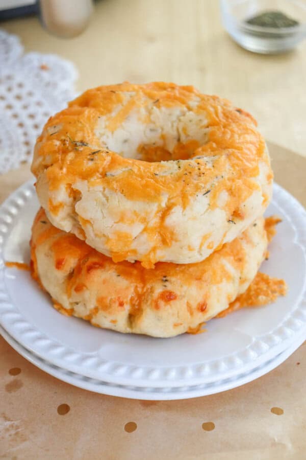 Two homemade cheddar cheese bagels stacked on a white plate. The bagels are golden brown with melted cheese on top, displayed on a light wooden surface with a doily in the background.