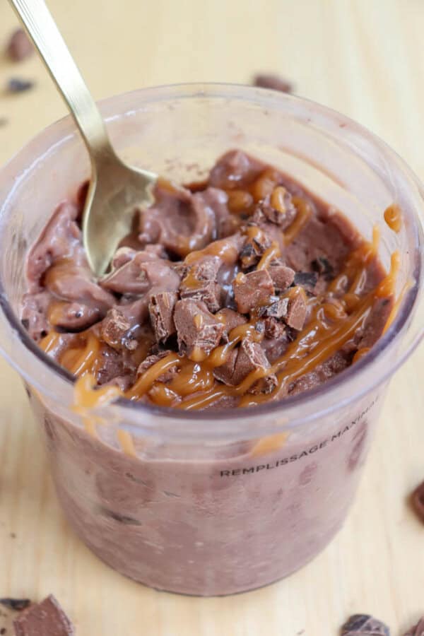 A clear cup filled with chocolate mousse, topped with caramel drizzle and chunks of chocolate. A golden spoon rests inside the mousse. The cup is placed on a wooden surface.