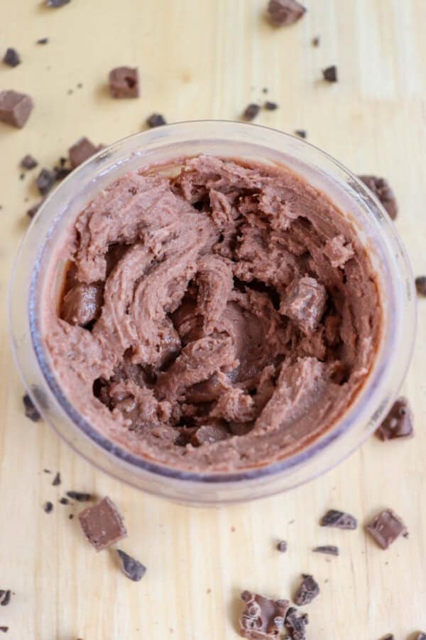 A glass bowl filled with creamy chocolate mousse, with chocolate chunks mixed in. The bowl is on a light wooden surface scattered with small pieces of chocolate.