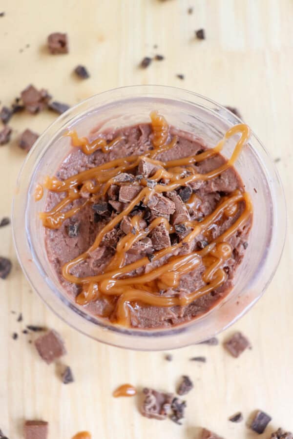 A plastic pint filled with chocolate mousse, topped with chopped chocolate pieces and drizzled with caramel sauce. The wooden surface around the cup has scattered chocolate bits.