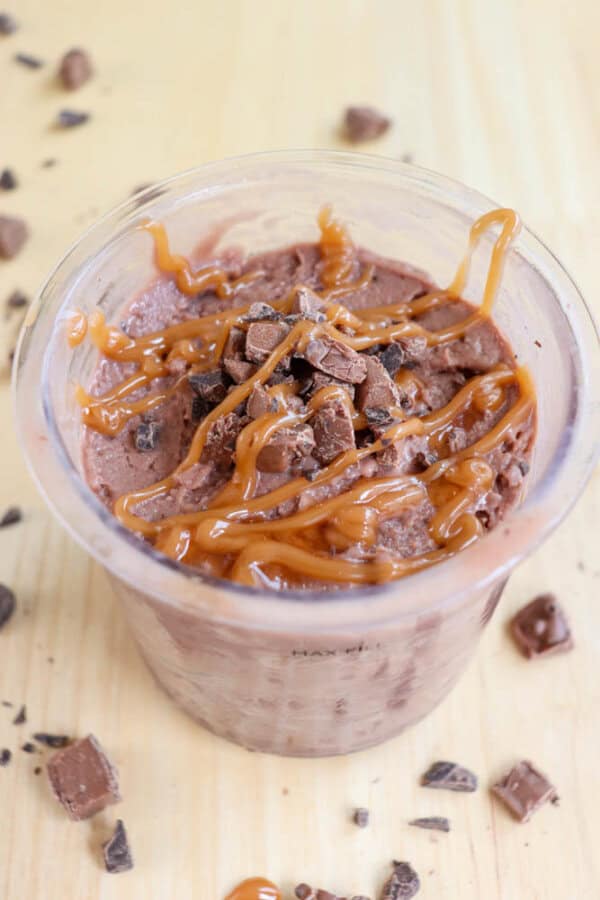 A creamy chocolate dessert topped with crushed chocolate pieces and caramel drizzle in a clear cup. Some chocolate chunks are scattered on the light wooden surface around the cup.