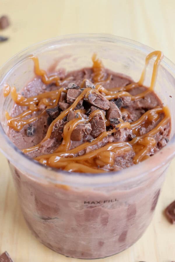 A close-up of a dessert in a clear cup, featuring chocolate ice cream mixed with chunks of chocolate. The top is drizzled with caramel sauce. The background is a light wooden surface with a few chocolate pieces scattered around.