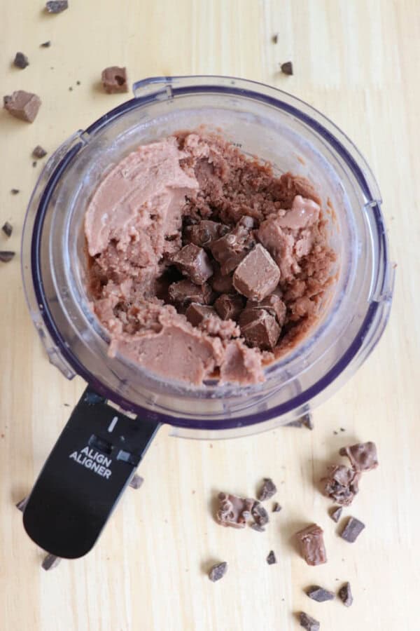 A small food processor containing chocolate ice cream mixed with chocolate chunks. The ice cream is partially scooped out, and additional chocolate pieces are scattered on the wooden surface around the processor.