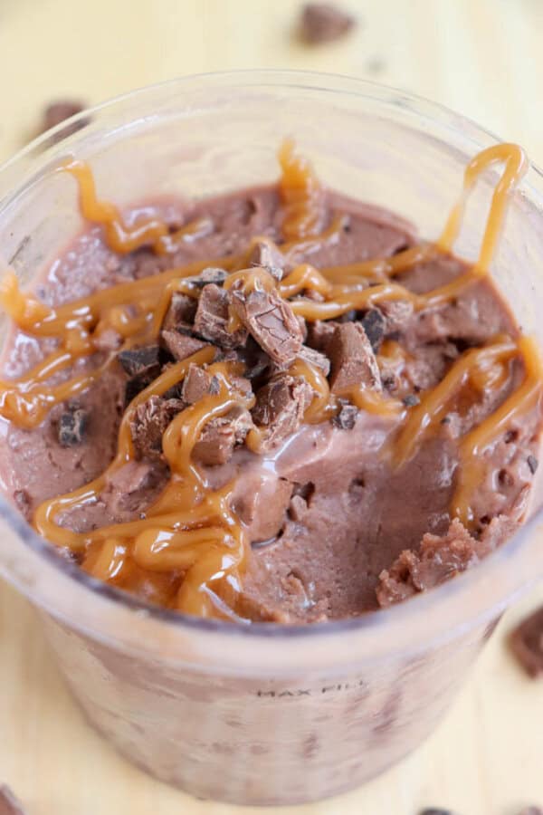 A close-up of a chocolate milkshake topped with chocolate chunks and drizzled with caramel sauce in a clear plastic cup.