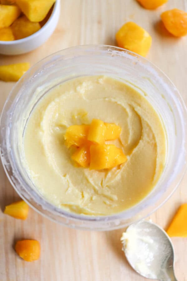 A bowl of creamy yellow mango frozen yogurt topped with small peach chunks, surrounded by a few scattered peach pieces on a wooden surface. A silver spoon lies beside the bowl.