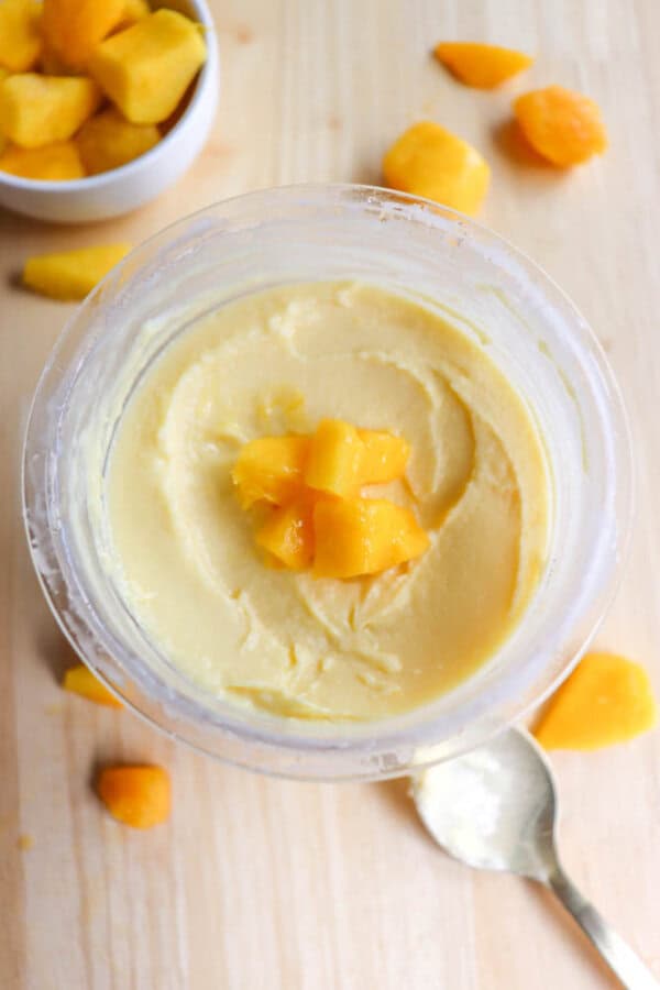 A glass jar filled with creamy yellow mango butter topped with small mango chunks. A spoon rests beside it on a light wooden surface, with a bowl of diced mango in the background and additional mango pieces scattered around.