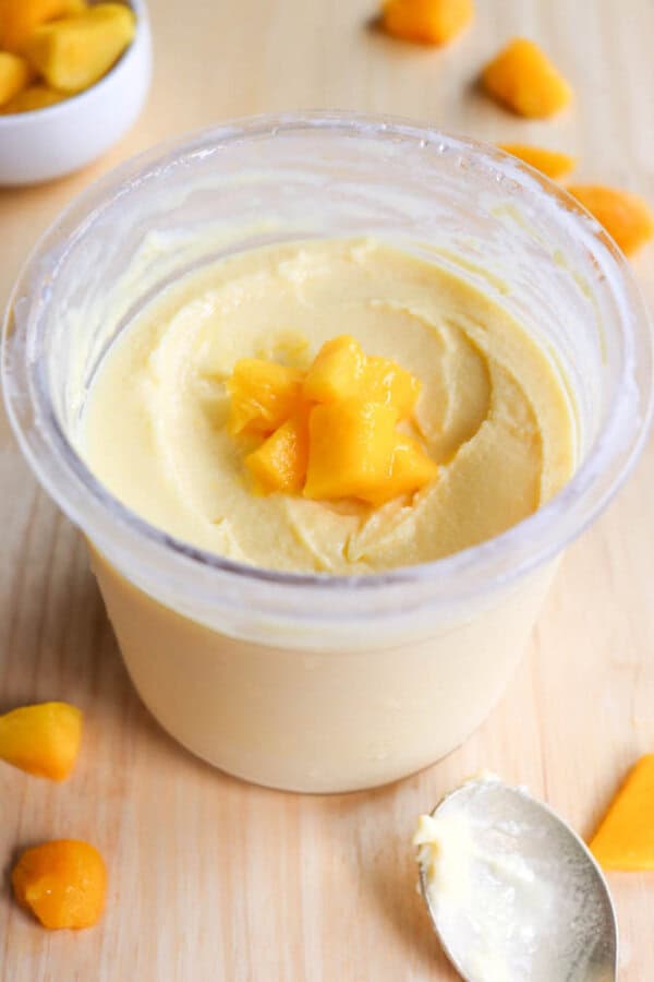 A clear container filled with creamy yellow mango ice cream, topped with small mango chunks, is placed on a wooden surface. There are scattered pieces of mango and a metal spoon nearby.