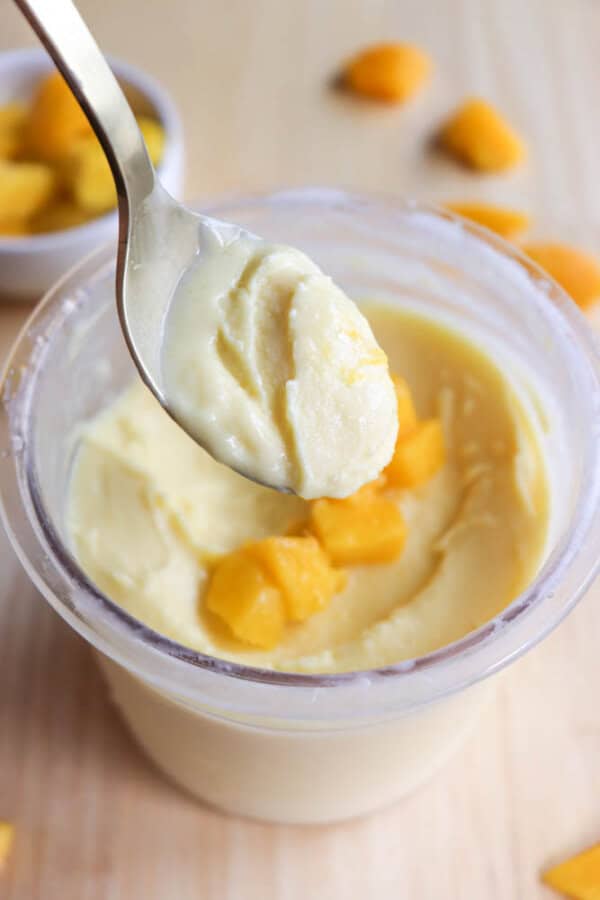 A spoon holds a creamy yellow dessert above an open container filled with the same dessert and topped with small mango chunks. Pieces of mango are scattered on the wooden surface in the background.