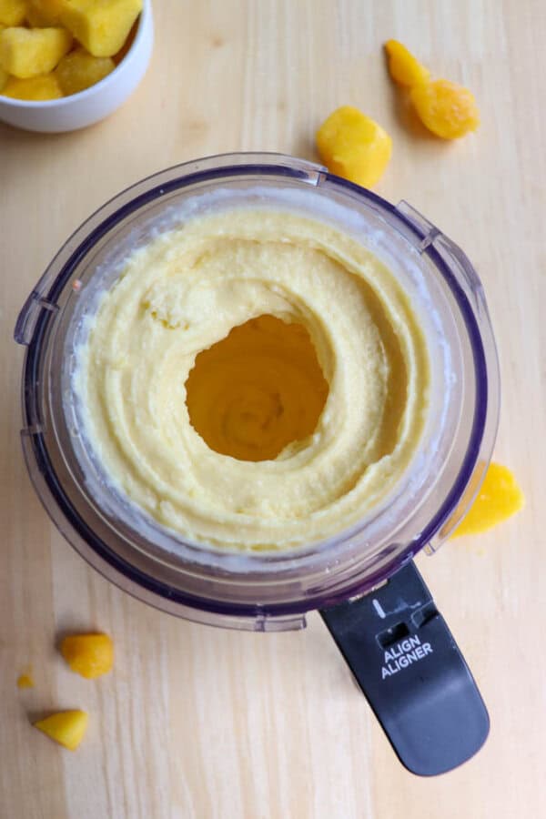 Top view of a food processor containing smooth, blended yellow mixture, likely mango puree. Small chunks of mango are scattered on the light wooden surface, with a small bowl of diced mango beside the processor.