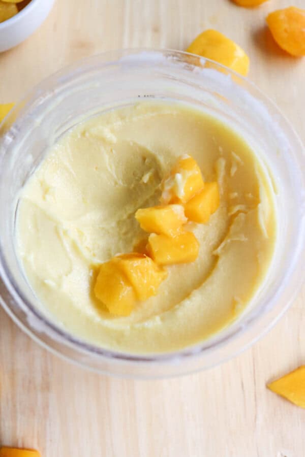 A bowl of creamy yellow mango mousse topped with chunks of fresh mango. The mousse sits on a light wooden surface, with additional mango pieces scattered around.