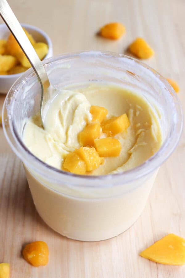 A clear cup filled with creamy mango frozen yogurt is topped with small cubes of fresh mango. A metal spoon is placed inside. Additional mango pieces are scattered on the wooden surface around the cup.