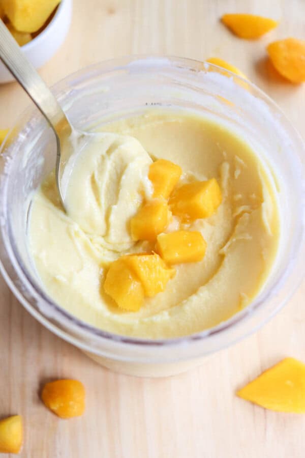 A spoon scoops creamy yellow dessert topped with small mango chunks in a clear bowl. Additional mango pieces are scattered on the light wooden surface around the bowl.