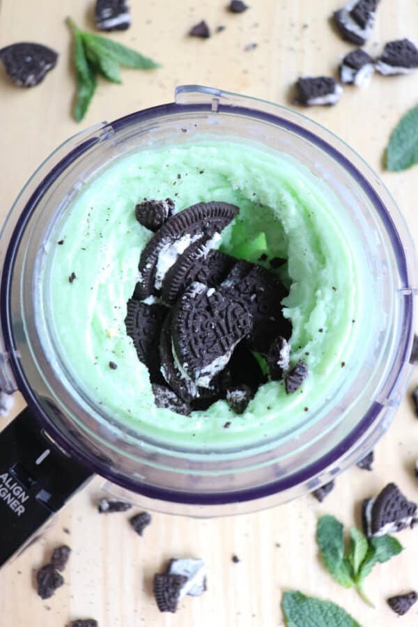 A ninja creami cup with mint green ice cream and Oreo cookies inside. Crushed Oreo pieces are scattered around the blender on the wooden surface, along with fresh mint leaves.