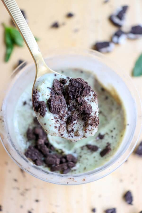 A spoonful of mint Oreo ice cream topped with crushed chocolate cookies, held above a bowl. More cookie crumbs are scattered on the wooden surface in the background.
