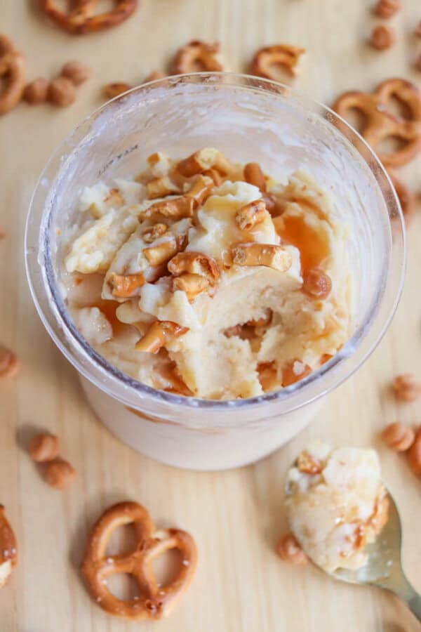 A glass jar filled with creamy ice cream topped with crushed pretzels and caramel sauce, placed on a wooden surface. Pretzel pieces and caramel bits are scattered around, and a spoon with ice cream rests nearby.