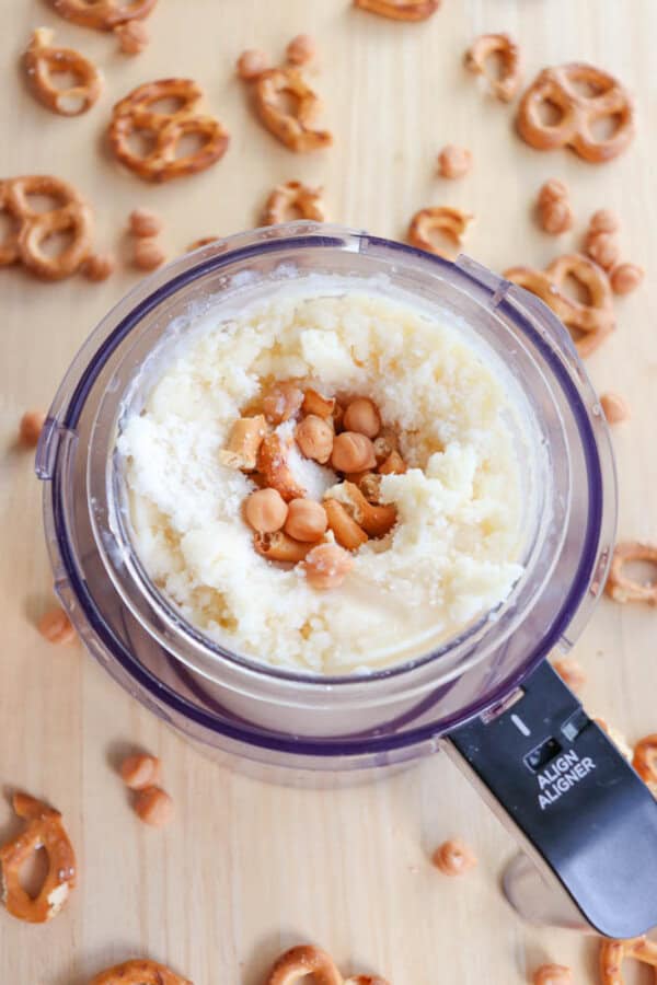 A food processor containing a mixture of crumbled pretzels and butterscotch chips is set on a wooden surface. Surrounding the processor are scattered pretzel pieces and butterscotch chips, creating a messy, inviting scene.